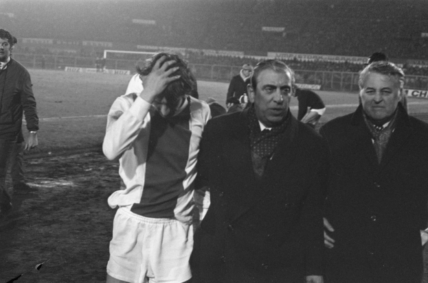 Schwarzes und weißes Bild einer Gruppe von Männern auf einem Fußballfeld, die enttäuscht nach einem Spiel aussehen, mit einem Stadion im Hintergrund.