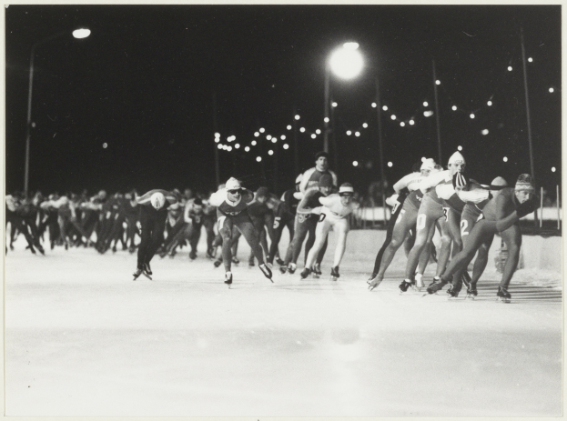 Eine Gruppe von Menschen, die nachts auf einer Eisbahn Schlittschuh laufen, im Hintergrund von Straßenlaternen beleuchtet, auf einem Schwarz-Weiß-Foto.