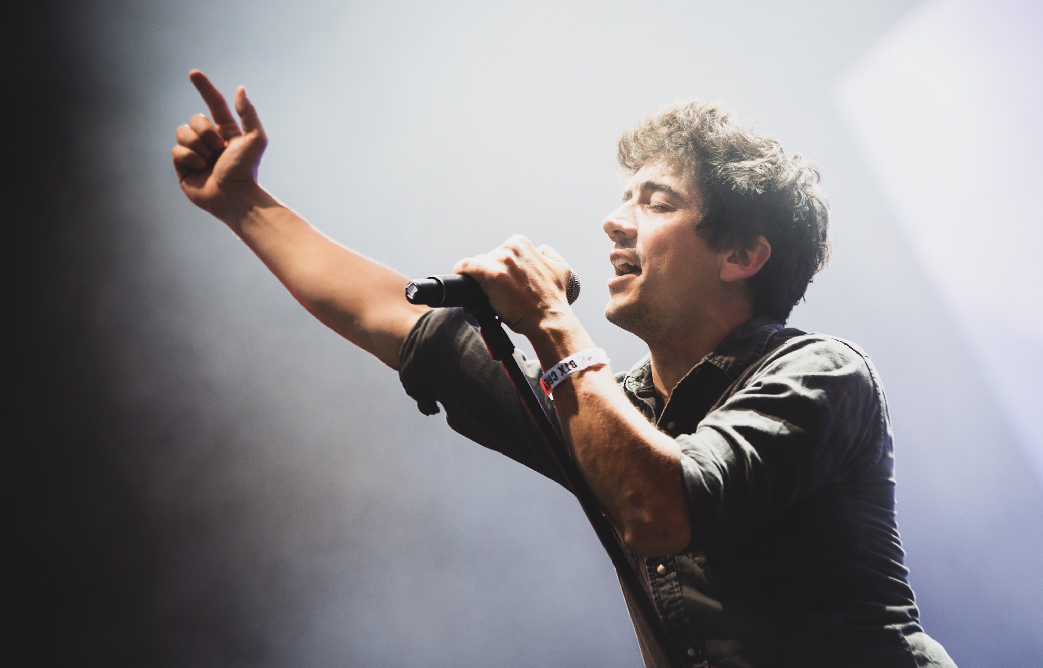 Ein Mann in schwarzem Hemd und Jeans singt leidenschaftlich in ein Mikrofon auf einer hell erleuchteten Bühne, mit geschlossenen Augen und stylisch verwuschelten Haaren, w├Ąhrend das Publikum im Hintergrund applaudiert.