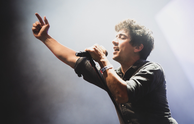 Ein Mann in schwarzem Hemd und Jeans singt leidenschaftlich in ein Mikrofon auf einer hell erleuchteten Bühne, mit geschlossenen Augen und stylisch verwuschelten Haaren, w├Ąhrend das Publikum im Hintergrund applaudiert.