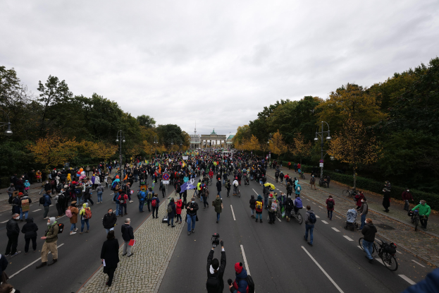 Eine große Gruppe von Menschen, die eine von Bäumen gesäumte Straße mit Laternenpfählen entlanggehen, halten dabei Kameras während einer Protestdemo in Berlin, mit einem Gebäude und einem klaren Himmel im Hintergrund.