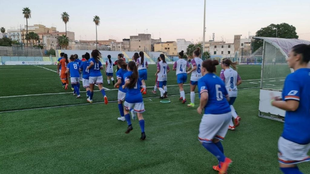 Eine Gruppe von Frauen, die auf einem Outdoor-Feld mit einem Tor und umgeben von Gebäuden, Bäumen und Pfählen unter einem klaren blauen Himmel Fußball spielen.