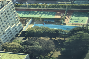 Eine Luftaufnahme eines Tennisplatzes in einer Stadt, umgeben von Gebäuden, Bäumen, Pflanzen, Pfählen, Fahrzeugen auf der Straße und einem Schwimmbad.