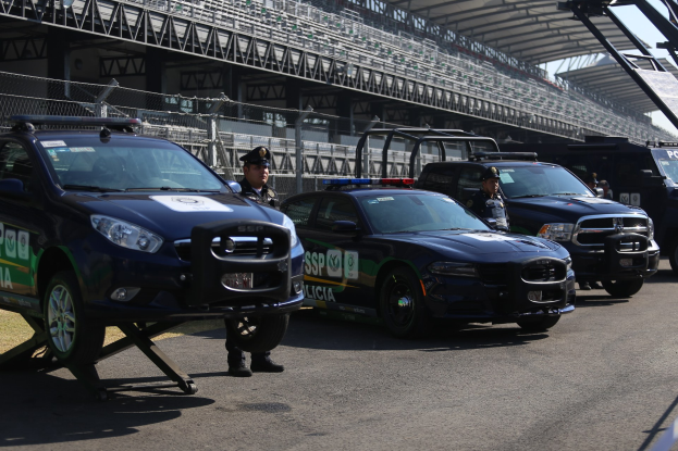 Eine Gruppe von Polizeiwagen vor einer Rennstrecke geparkt, mit uniformierten Beamten auf der Straße stehend, im Hintergrund ein Stadion und Maschendrahtzaun.