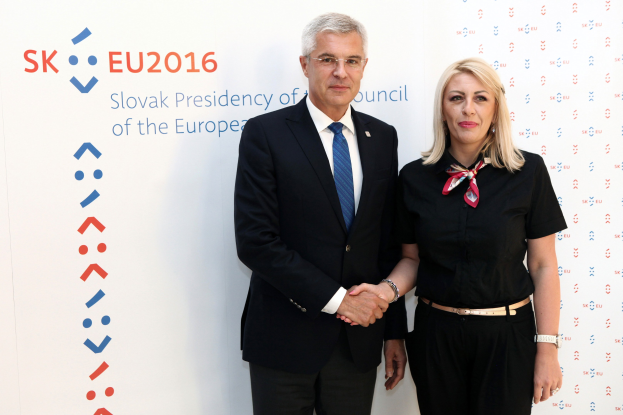 Zwei Personen, ein Mann in einem schwarzen Blazer und weißem Hemd mit blauer Krawatte und eine Frau mit Brille, stehen lachend vor einer weißen Tafel mit "sk eu 2016" geschrieben, was darauf hinweist, dass sie der Präsident des Europäischen Rates sind.