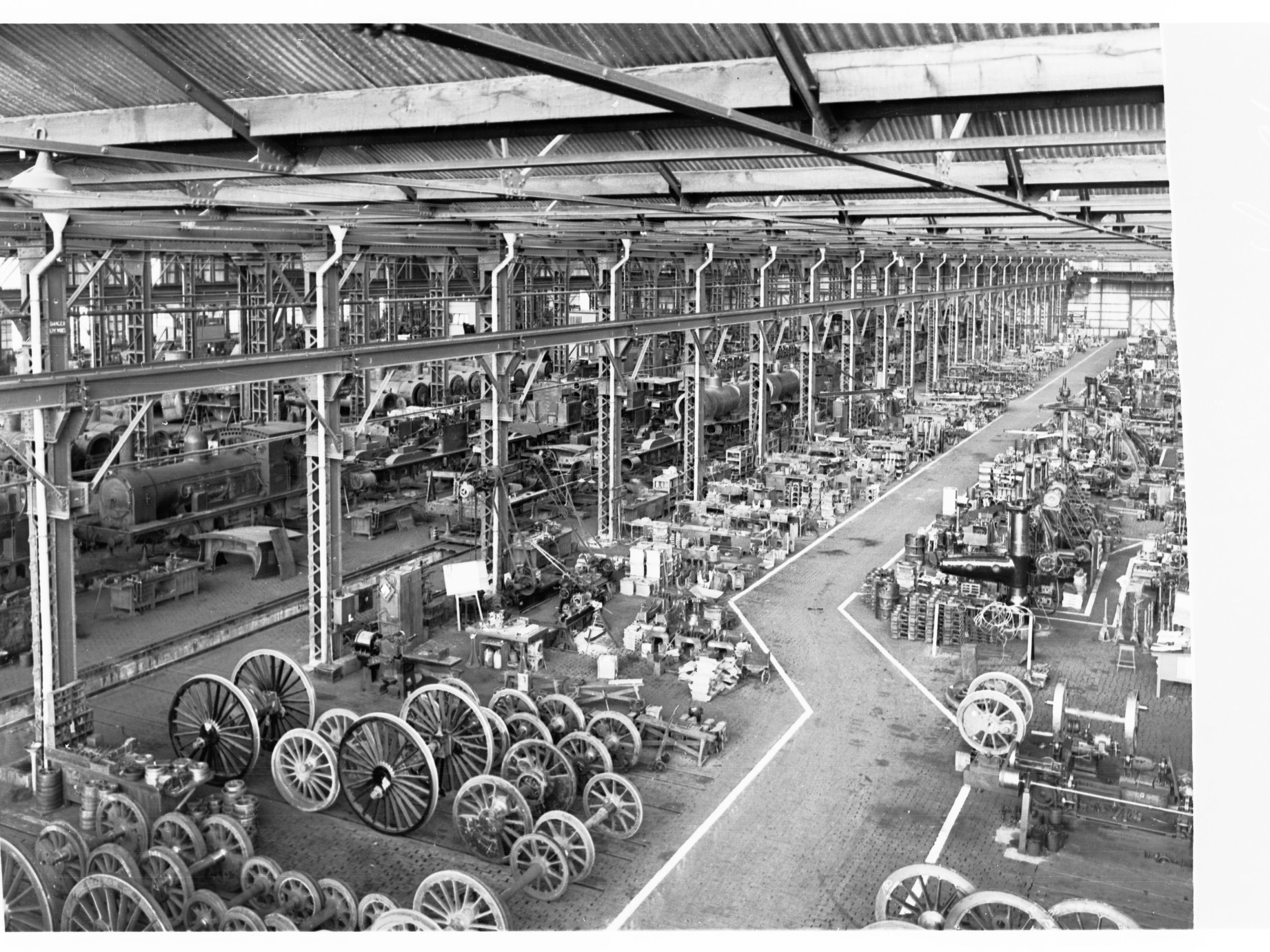 Ein Schwarz-Weiß-Foto eines Fabrikinneren, das mit verschiedenen Maschinen, einschließlich Rädern, Maschinen und Pfählen, gefüllt ist, mit einem Schuppen oben.