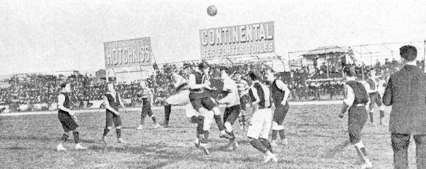Ein Schwarz-Weiß-Bild von einer Gruppe von Männern, die auf einem Feld Fußball spielen, mit einem Ball in der Luft und Zuschauern im Hintergrund, mit Tafeln, die wahrscheinlich textbezogen sind.