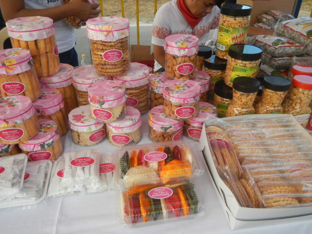 Eine Gruppe von Menschen steht um einen Tisch mit verschiedenen Lebensmitteln an einem Food-Festival, im Hintergrund ist ein Geländer und eine Straße zu sehen.