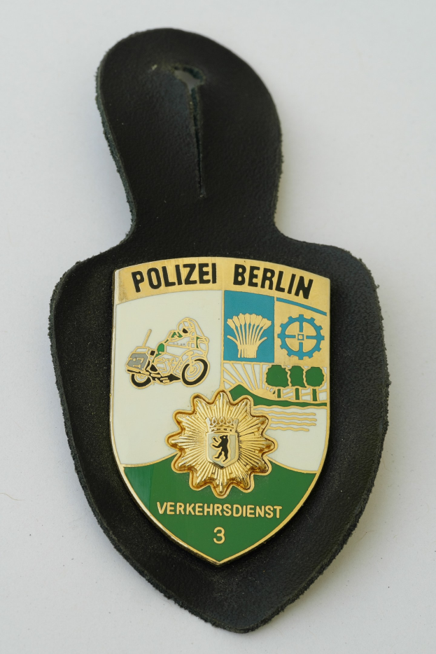 Ein schildförmiges Polizeibadges mit "Polizei Berlin" in schwarzer fetter Schrift auf einem blauen Hintergrund mit einem weißen Stern und Rahmen, das auf einer Oberfläche ruht.