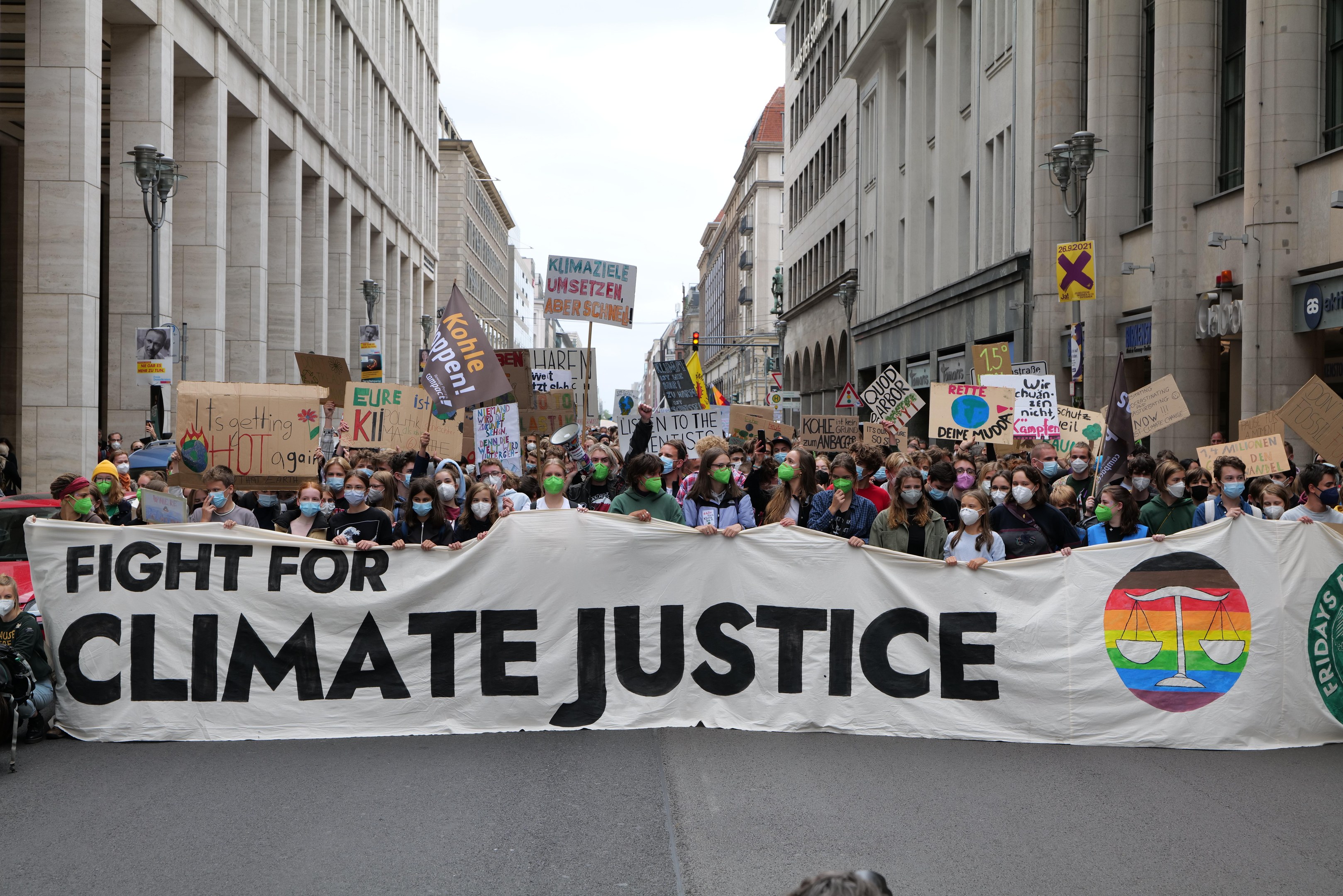 Eine große Gruppe von Menschen marschiert durch eine Stadtstraße und hält ein 'Kampf für Klimagerechtigkeit'-Schild und trägt Masken, mit Gebäuden auf beiden Seiten und einem klaren blauen Himmel im Hintergrund.
