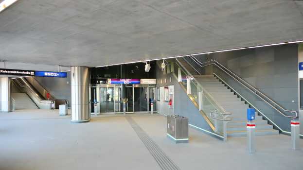 Leerer Bahnhof mit Treppe, Geländern, bodennahen Mülltonnen, Deckenleuchten, Texttafeln und Glastüren im Hintergrund.