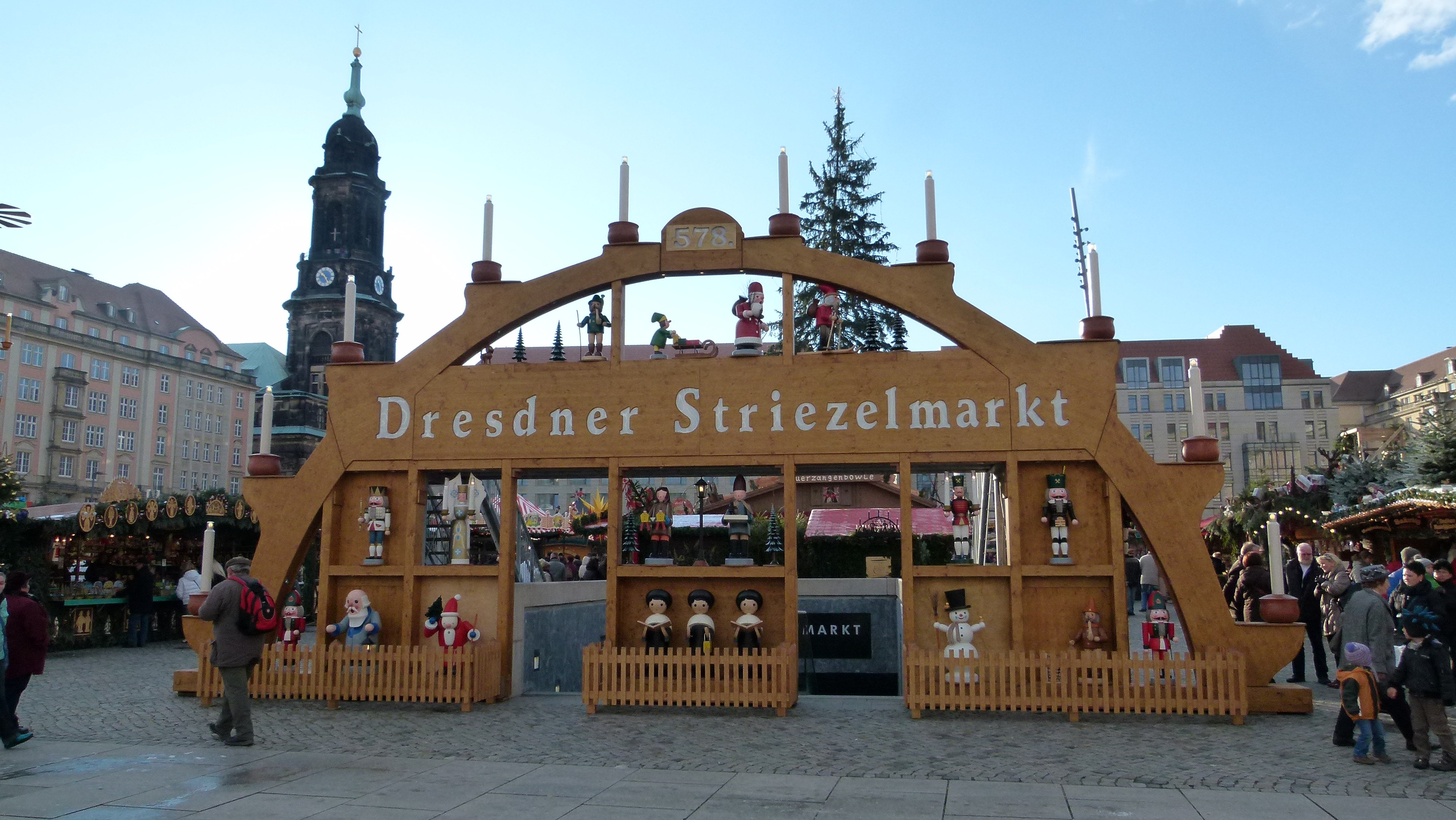 Ein lebendiger Weihnachtsmarkt in Dresden, Deutschland, mit Menschen, die herumlaufen, einige mit Taschen, ein Bogen mit Text, Bäume, Gebäude mit Fenstern und ein bewölkter Himmel.