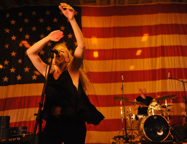 Eine Frau singt in ein Mikrofon auf der Bühne mit einer amerikanischen Flagge im Hintergrund, begleitet von einem Schlagzeuger, mit Bühnenausrüstung und Beleuchtung im Sichtfeld.
