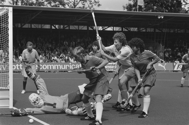 Schwarzes Bild einer Gruppe von Frauen, die Feldhockey spielen, Helme tragen und Stöcke halten, mit einem Tor auf der linken Seite, Zuschauern in den Rängen, Bannern, einem Schuppen, Bäumen und einer Flagge im Hintergrund.
