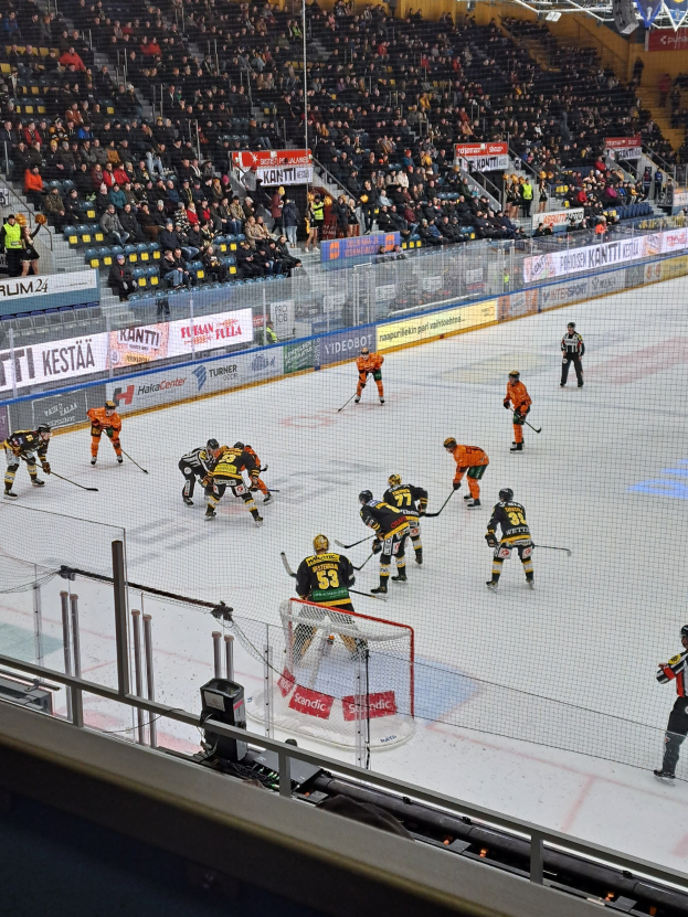 Eine Gruppe von Menschen, die Hockey auf einer Indoor-Eisbahn spielen, umgeben von einem Zaun, mit Zuschauern im Hintergrund und beleuchteter Stadionbeleuchtung oben.
