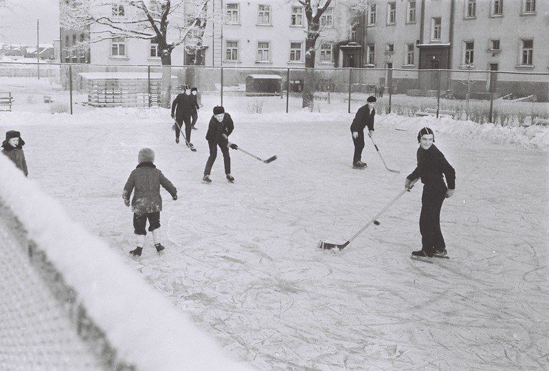 Schwarzes und weißes Bild von Menschen, die Hockey auf einem Eisplatz spielen, der von einem Zaun, Bäumen, Gebäuden mit Fenstern und einer Bank umgeben ist.