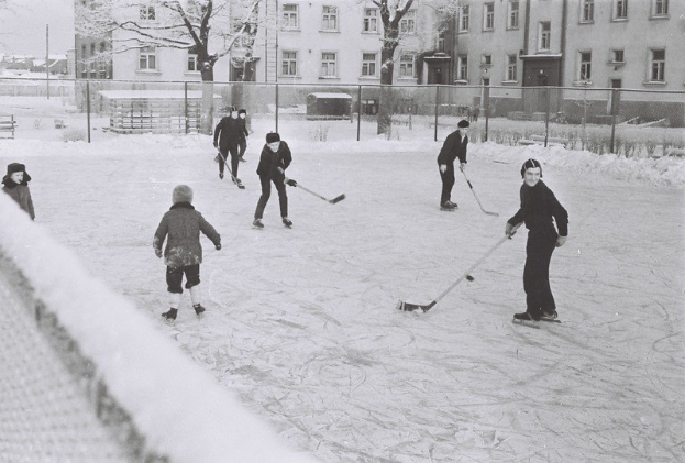 Schwarzes und weißes Bild von Menschen, die Hockey auf einem Eisplatz spielen, der von einem Zaun, Bäumen, Gebäuden mit Fenstern und einer Bank umgeben ist.