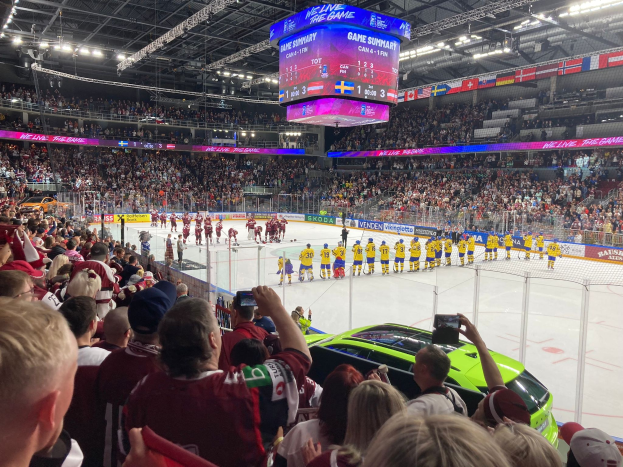 Ein Eishockeyspiel in einer großen Arena mit Spielern auf dem Eis, Zuschauern auf Sitzen und Stehplätzen mit Kameras, einem Netz in der Mitte und Lampen und einem Bildschirm oben.