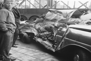 Ein Schwarz-Weiß-Bild eines verunglückten Autos am Straßenrand mit mehreren Personen drumherum, einem Lkw und einer überbrückung im Hintergrund.