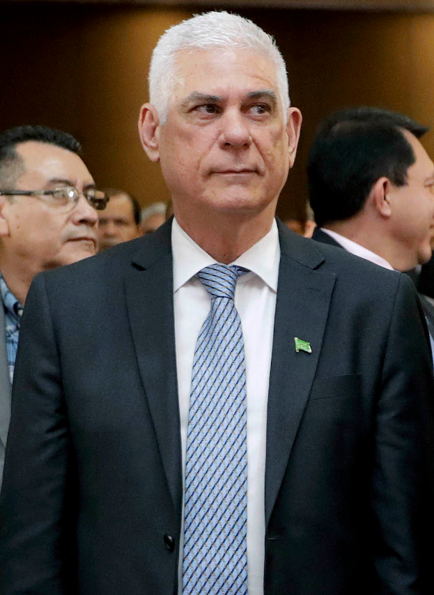 Ein Mann in einem blauen Anzug, weißem Hemd und gemustertem Schlips steht vor einer Gruppe von Menschen und hat einen ernsten Gesichtsausdruck und ordentliches Haar.