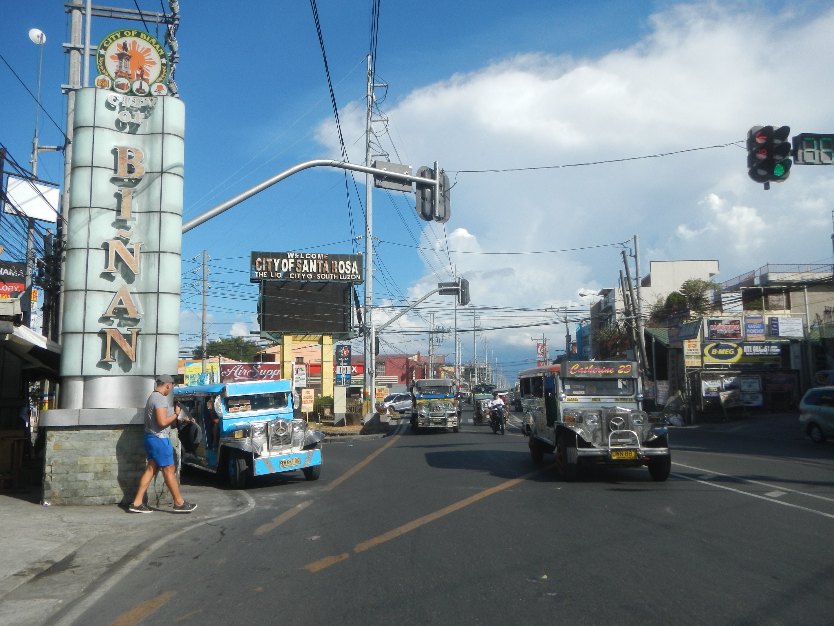 Eine belebte Stadtstraße mit Fahrzeugen, Fußgängern, Ampeln, Gebäuden, Bäumen und Oberleitungen.