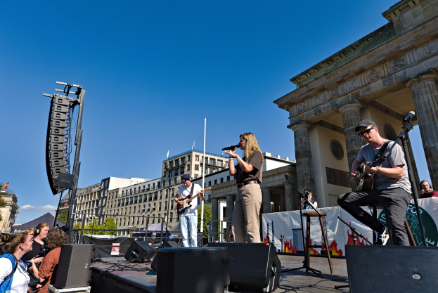 Eine Gruppe von Menschen, die auf einer Bühne vor dem Brandenburger Tor in Berlin Musik machen, umgeben von Lautsprechern und Equipment, vor einem klaren blauen Himmel.