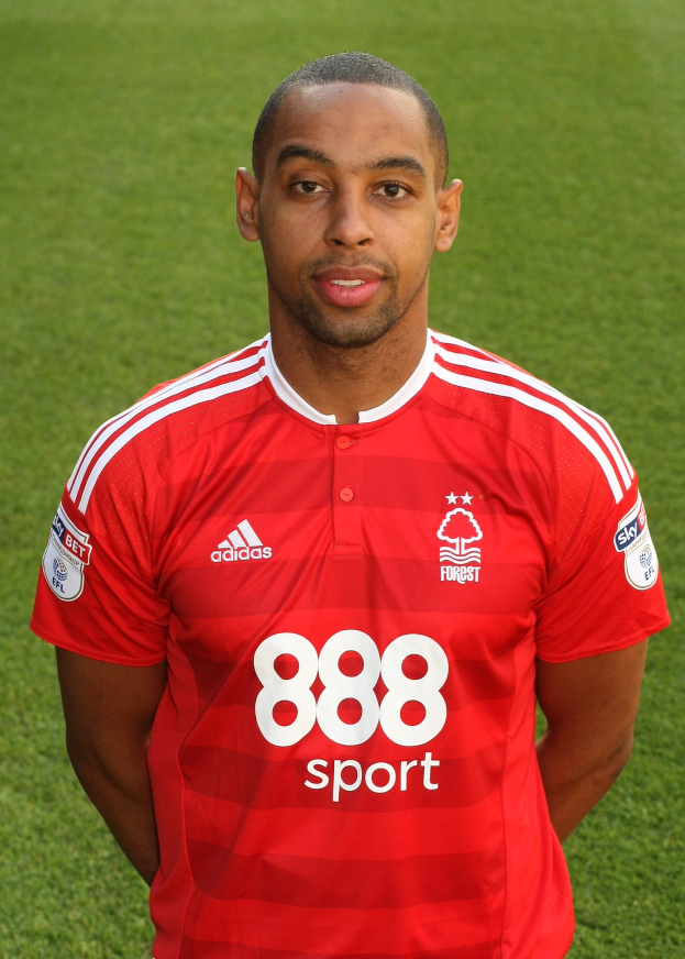 Ein Mann in einem roten T-Shirt steht selbstbewusst auf einem Fußballfeld und sieht aus wie ein Flügelspieler mit entschlossener Miene.