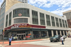 Eine belebte Stadtstraße mit fahrenden Fahrzeugen und Fußgängern auf dem Gehweg, hohe Glasgebäude im Hintergrund und ein Schild mit der Aufschrift "The Best Value Urban Outlet" unter einem bewölkten Himmel.