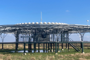 Große metallene Solarstromanlage mit Treppe, Geländer und umgebenen Pfählen mit Lampen, in einem grasbewachsenen Feld mit Wasser, Bäumen und einem bewölkten Himmel im Hintergrund.