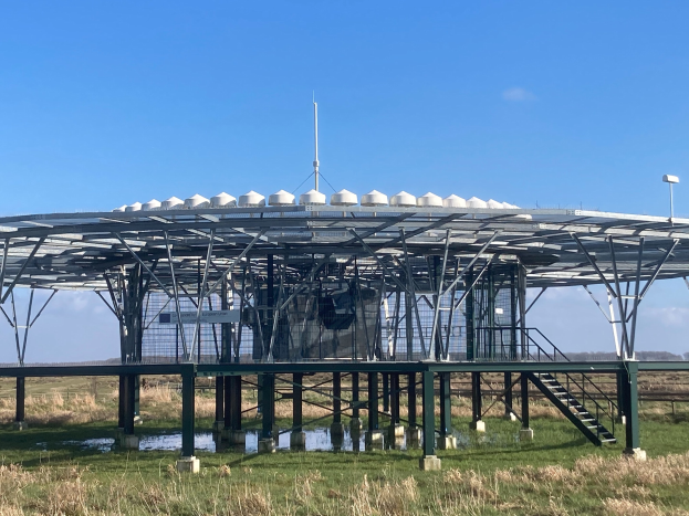 Große metallene Solarstromanlage mit Treppe, Geländer und umgebenen Pfählen mit Lampen, in einem grasbewachsenen Feld mit Wasser, Bäumen und einem bewölkten Himmel im Hintergrund.
