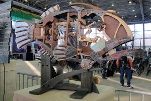 Große Metallfischskulptur in einem Innenraum mit Zuschauern, umgeben von Stühlen, einem Zaun, einer Fahne, einem Lautsprecher, Vorhängen und Deckenleuchten.