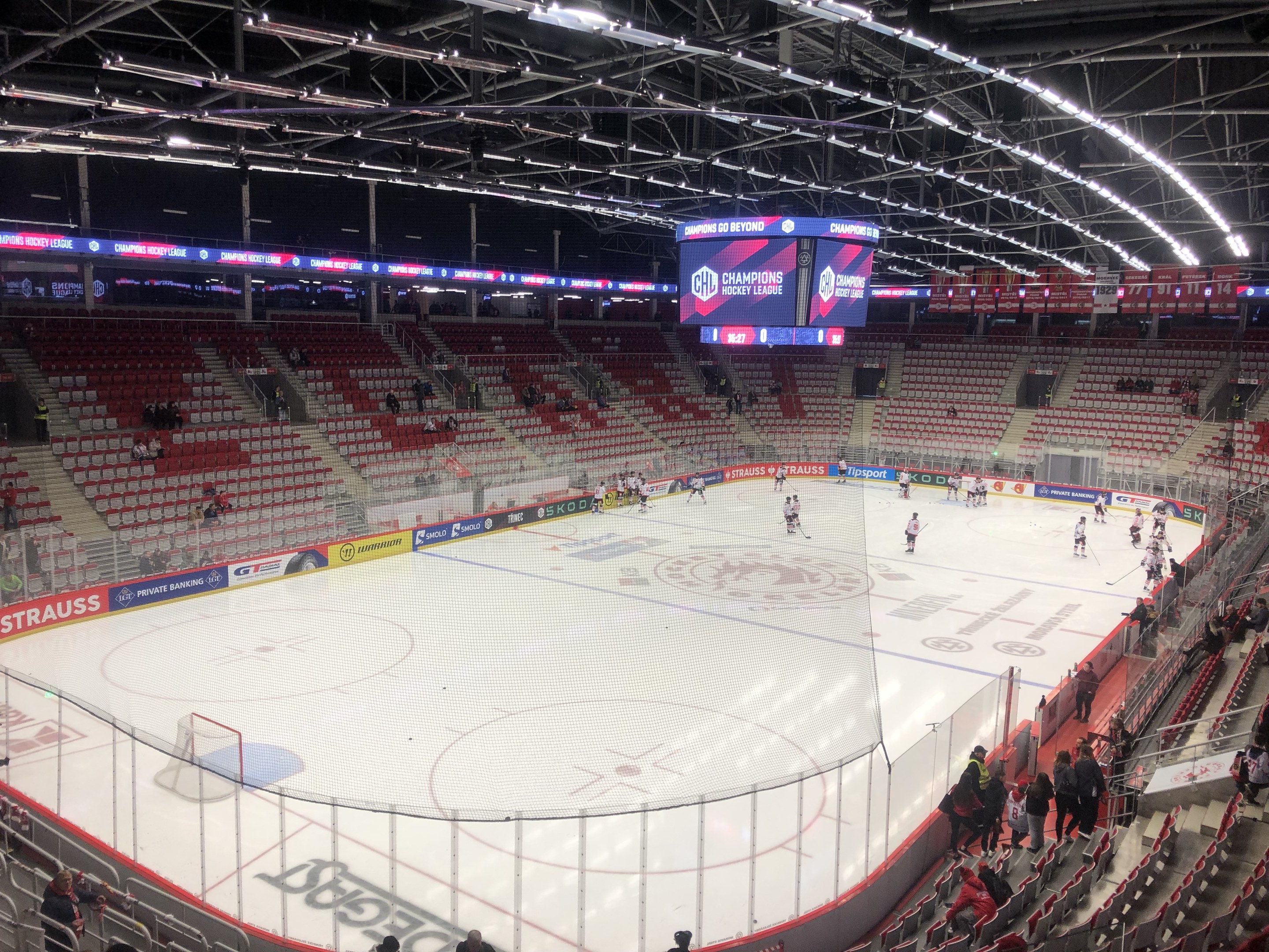 Eishockeyspiel in einer großen Arena zwischen den St. Louis Blues und Detroit Red Wings, mit Zuschauern auf den Sitzplätzen und Stehplätzen in der Nähe der Eisfläche umgeben von einem Zaun und Metallrahmen, unter einem Dach mit Deckenlampen und einem Anzeigebildschirm.