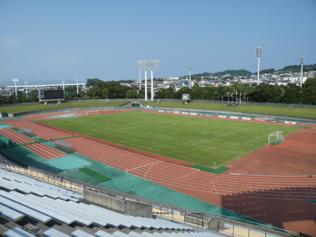 Großes Stadion mit einem Fußballfeld, umgeben von grünem Gras, Bäumen, Laternenmasten, einigen Menschen, leeren Stühlen, Gebäuden und einem klaren blauen Himmel.