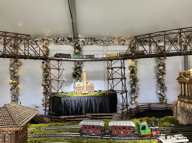 Eine Modellbahnanlage auf einer Schiene in einem Zelt, umgeben von Miniaturhäusern, Schienen, einer Brücke, dekorativen Gegenständen und anderen Objekten, mit einer Wand im Hintergrund, die eine festliche Weihnachtsbahnschau-Atmosphäre schafft.
