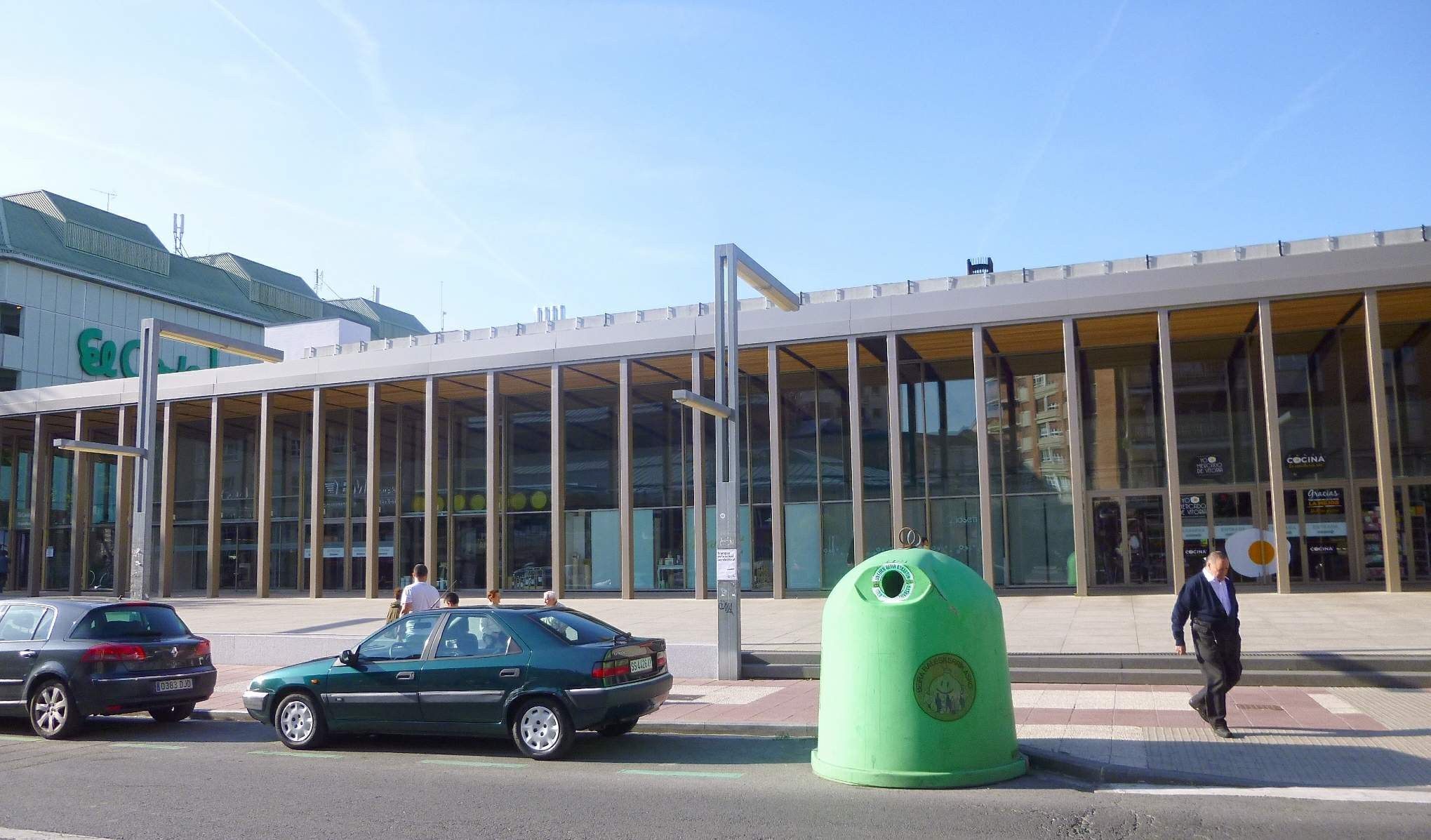 Grüner Müllcontainer auf der Straße neben einem Gebäude mit Glaswänden und Säulen, mit Fahrzeugen und Fußgängern im Vordergrund und einem bewölkten Himmel im Hintergrund.