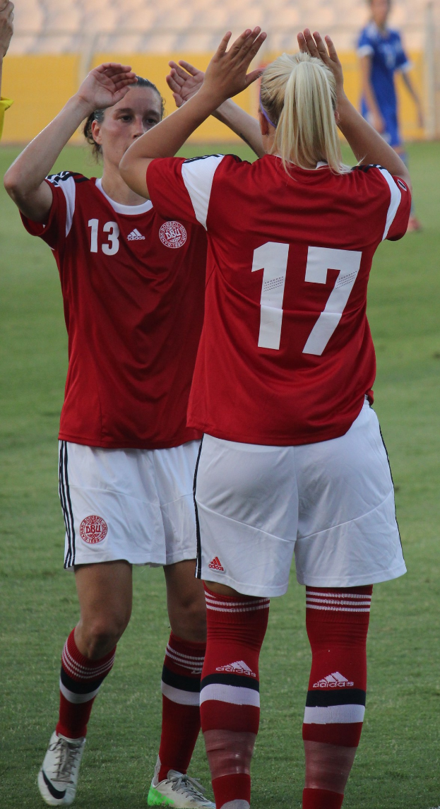 Zwei Frauen in roten T-Shirts und weißen Shorts klatschen auf einem Fußballfeld mit einer Person in einem blauen Kleid und einem gelben Board im Hintergrund.