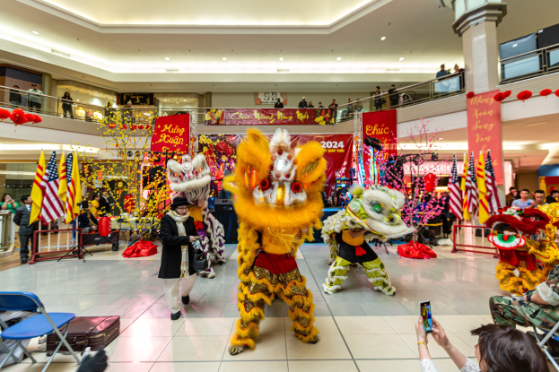Ein lebhaftes chinesisches Neujahrsfest in einem Einkaufszentrum mit Menschen in Kostümen, die tanzen, andere auf Stühlen sitzen und Handys halten, umgeben von Flaggen, Bannern, Dekorationen, Geländern, Säulen und Deckenleuchten.