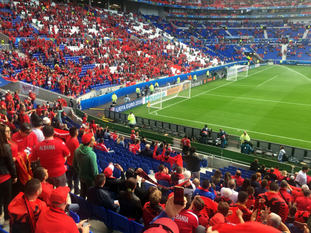 Ein volles Stadion mit Zuschauern, einige sitzen, andere stehen, mit Bannern, einem Tor und Deckenleuchten, die gespannt auf den Beginn eines Fußballspiels warten.