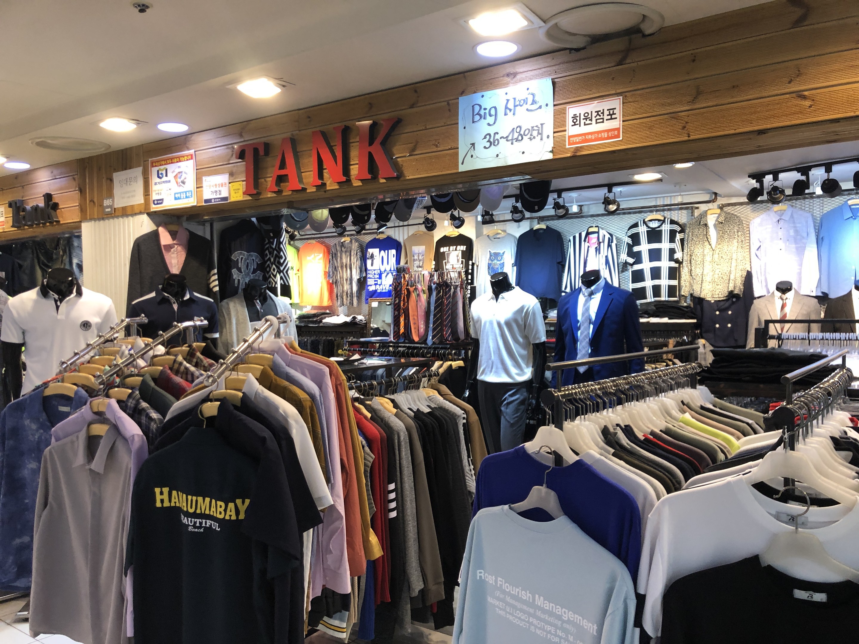 Ladeninneneinrichtung mit zahlreichen Hemden an der Wand, Schaufensterpuppen mit aktuellen Outfits, Texttafeln an den Wänden und moderner Deckenbeleuchtung.