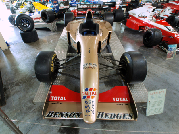 Eine Gruppe von Rennwagen in einem Motorsportmuseum, getrennt durch einen Metallzaun mit einer Texttafel links.