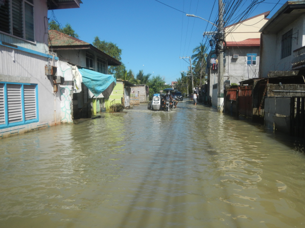 Eine überflutete Straße in einem kleinen Dorf mit Menschen, die durch das Wasser gehen, Gebäude mit Fenstern und Türen auf beiden Seiten, Strommasten mit Drähten, Bäume und einen klaren Himmel im Hintergrund.