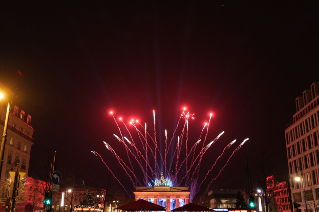 Eine nächtliche Stadtstraße mit dem beleuchteten Brandenburger Tor in Berlin, Deutschland, umgeben von Gebäuden, Bäumen, Laternen, Verkehrszeichen, Schildern, Zelten und Menschen, mit Feuerwerk am Himmel.