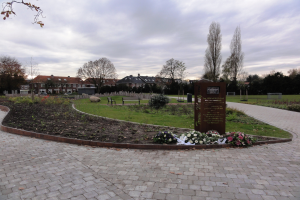 Eine Gedenkstätte im Park mit einem Backsteinweg, umgeben von Grün, Bänken und Bäumen, mit Gebäuden und einem bewölkten Himmel im Hintergrund und einer Tafel mit Text auf der rechten Seite.