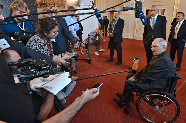 Ein Mann im Rollstuhl wird von Reportern mit Kameras, Handys und Mikrofonen mit Ständern umgeben, mit einem Banner, Scheinwerfern und einer Wand im Hintergrund.