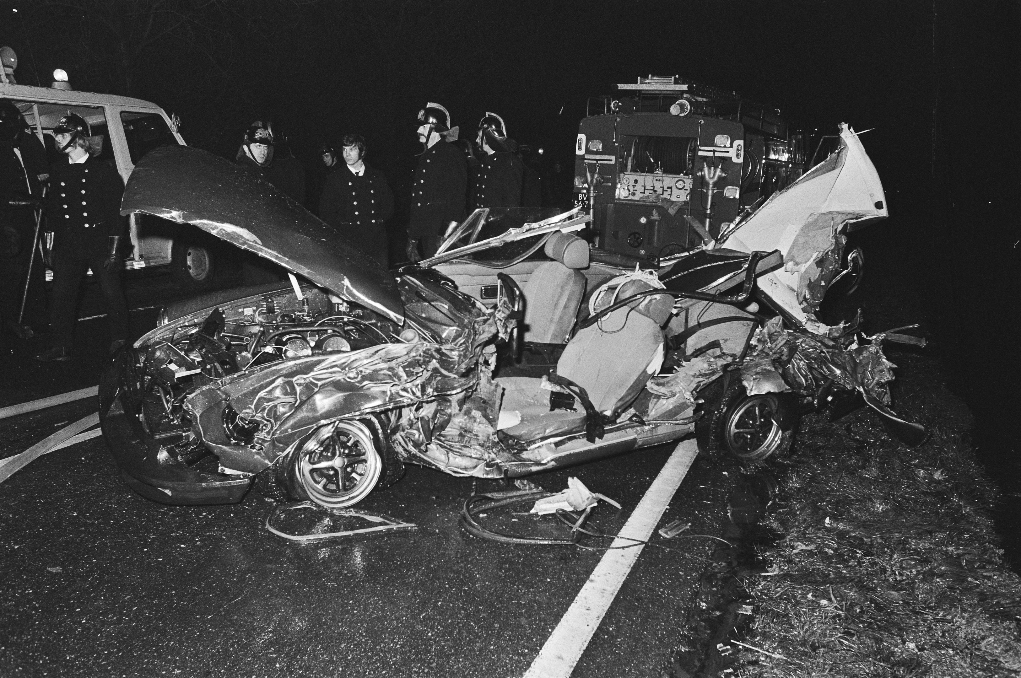 Schwarzes und weißes Foto eines beschädigten Autos am Straßenrand mit mehreren Personen, einige mit Helmen, die darum herum stehen und ein Fahrzeug im Hintergrund.