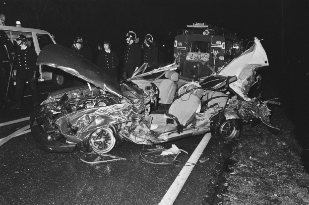 Schwarzes und weißes Foto eines beschädigten Autos am Straßenrand mit mehreren Personen, einige mit Helmen, die darum herum stehen und ein Fahrzeug im Hintergrund.