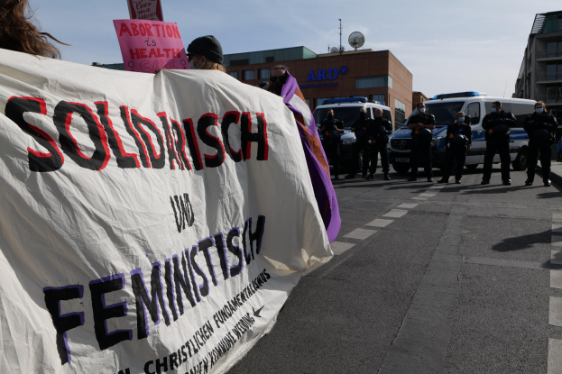 Eine Gruppe von Menschen marschiert auf einer Straße, eine Fahne haltend, auf der "Solidarität und Feminismus" steht, mit parkenden Fahrzeugen am Straßenrand und Gebäuden im Hintergrund unter einem klaren blauen Himmel.