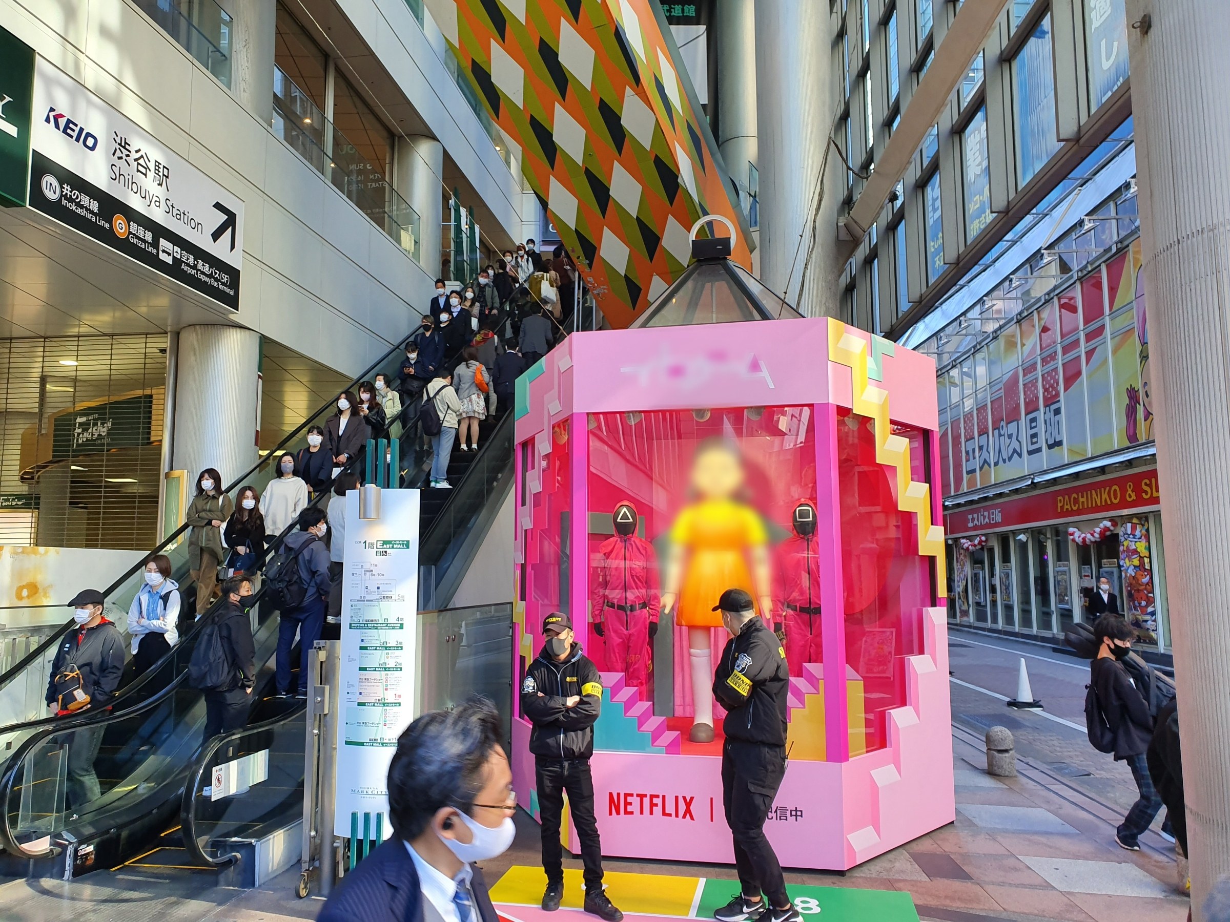 Eine Gruppe von Menschen steht vor einem Gebäude mit Glaswänden, Texttafeln, einige tragen Masken, in der Nähe einer Rolltreppe; eine pinkfarbene Box in der Mitte zeigt Schaufensterpuppen und trägt 'Netflix's neuen Netflix-Schalter in Tokyo.'