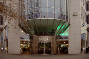 Großes Glasgebäude mit "BMW"-Text, Menschen vor dem Gebäude und einem Baum links.