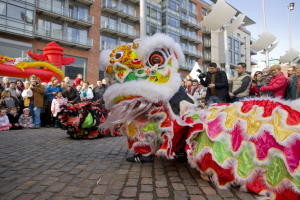 Ein lebendiges chinesisches Neujahrsfest in Amsterdam mit einem Löwen tanzen im Vordergrund, umgeben von einer Menschenmenge, einige halten Kameras, vor einem Hintergrund von Gebäuden, Laternenmasten und einem klaren blauen Himmel.